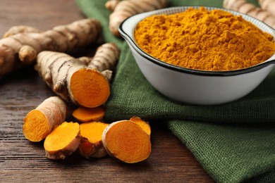 Photo of Bowl with aromatic turmeric powder and cut roots on wooden table, closeup