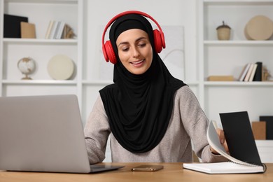 Muslim woman in headphones studying near laptop at wooden table in room