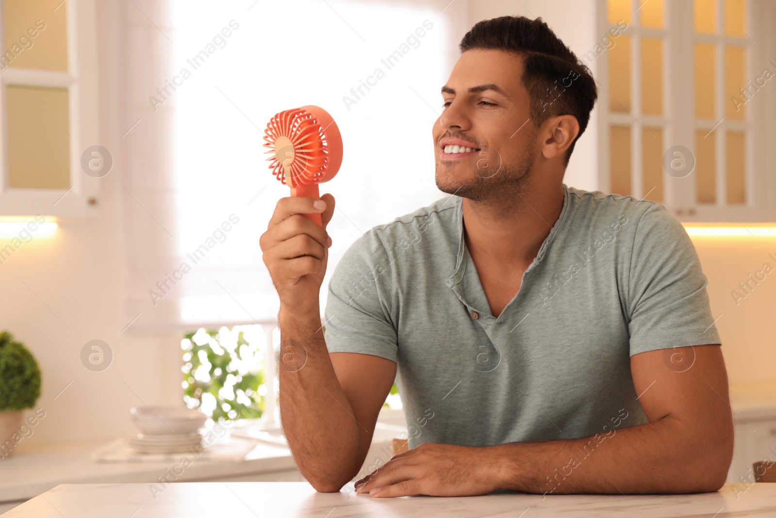 Photo of Man enjoying air flow from portable fan at table in kitchen. Summer heat