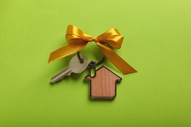 Key with trinket in shape of house and yellow bow on light green background, top view. Housewarming party