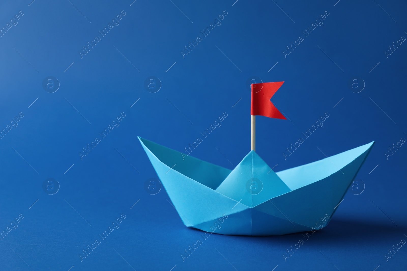 Photo of Handmade paper boat with red flag on blue background. Space for text. Origami art