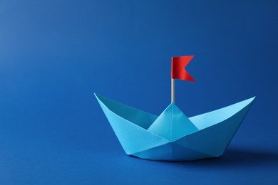 Photo of Handmade paper boat with red flag on blue background. Space for text. Origami art