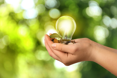 Image of Solar energy concept. Woman holding glowing light bulb with seedling and coins against green blurred background, closeup