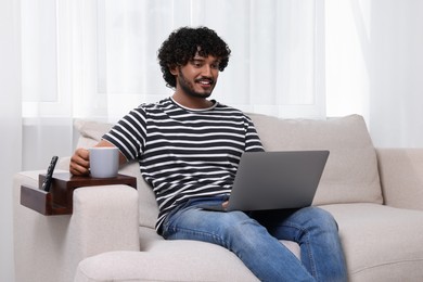 Photo of Happy man using laptop and holding cup of drink on sofa with wooden armrest table at home