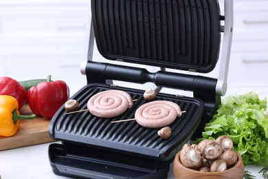 Photo of Electric grill with homemade sausages, mushrooms and vegetables on white table