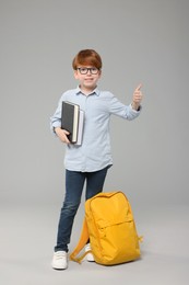Happy schoolboy with books showing thumb up on grey background