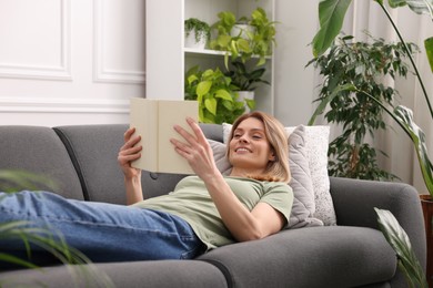 Photo of Woman reading book on sofa in room with houseplants