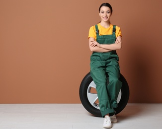 Female mechanic in uniform with car tire on color wall background