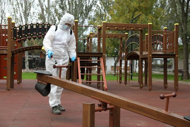 Photo of Woman wearing chemical protective suit with disinfectant sprayer on playground. Preventive measure during coronavirus pandemic