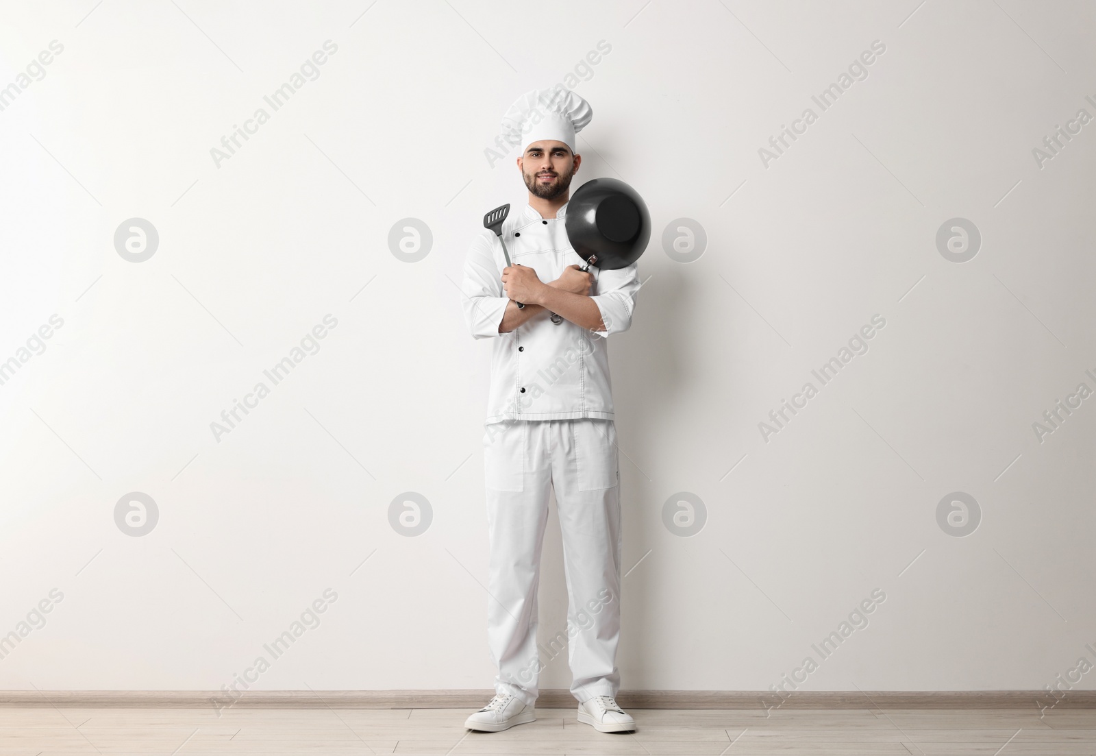 Photo of Professional chef with wok and spatula near white wall indoors