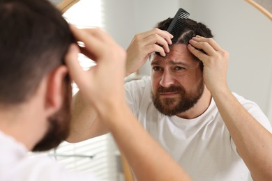 Photo of Dandruff problem. Man with comb examining his hair and scalp near mirror indoors