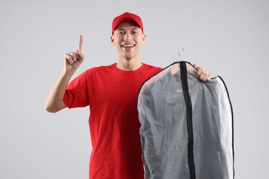 Photo of Dry-cleaning delivery. Happy courier holding garment cover with clothes and pointing at something on light grey background