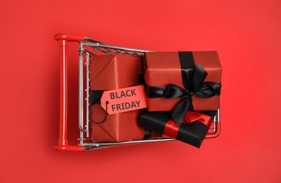 Photo of Small shopping cart with wrapped gift boxes on red background, top view. Black Friday sale