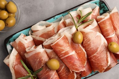 Photo of Rolled slices of delicious jamon with rosemary and olives on grey table, flat lay