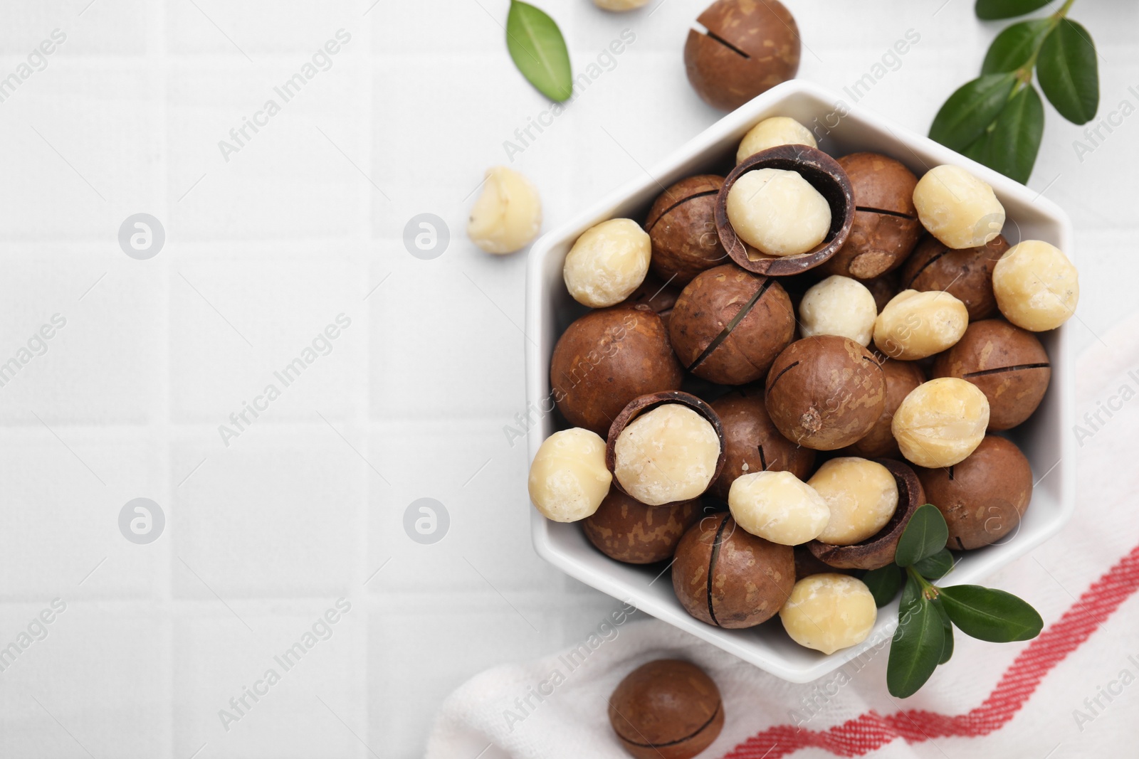 Photo of Tasty Macadamia nuts in bowl and green twigs on white tiled table, flat lay. Space for text