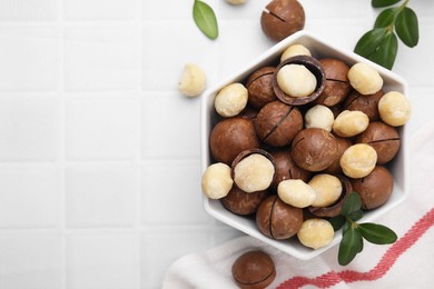 Photo of Tasty Macadamia nuts in bowl and green twigs on white tiled table, flat lay. Space for text