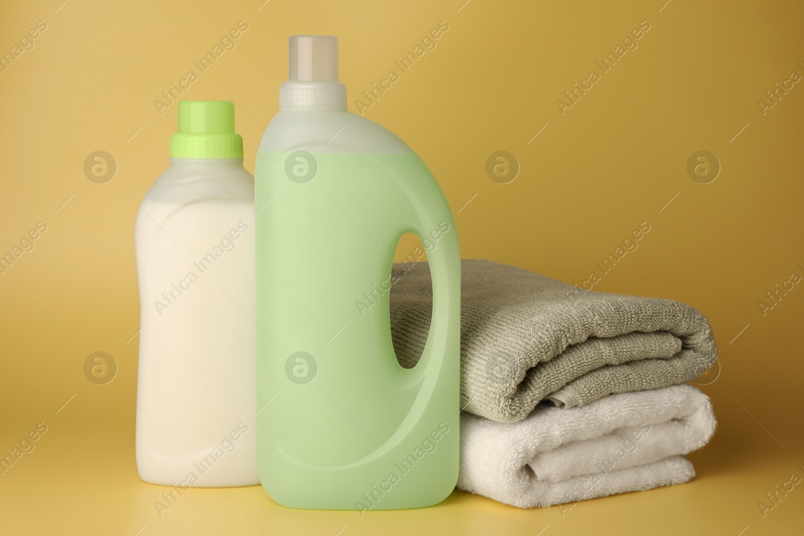 Photo of Bottles of fabric softener and stacked clean towels on pale yellow background