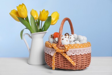Photo of Wicker basket with festively decorated Easter eggs and beautiful tulips on white wooden table against light blue background