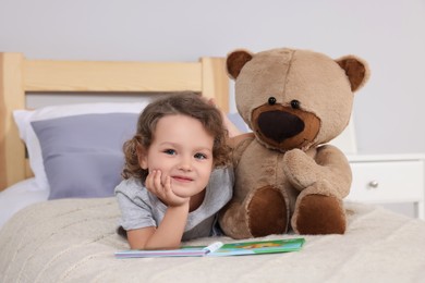 Cute little girl with book and teddy bear on bed at home