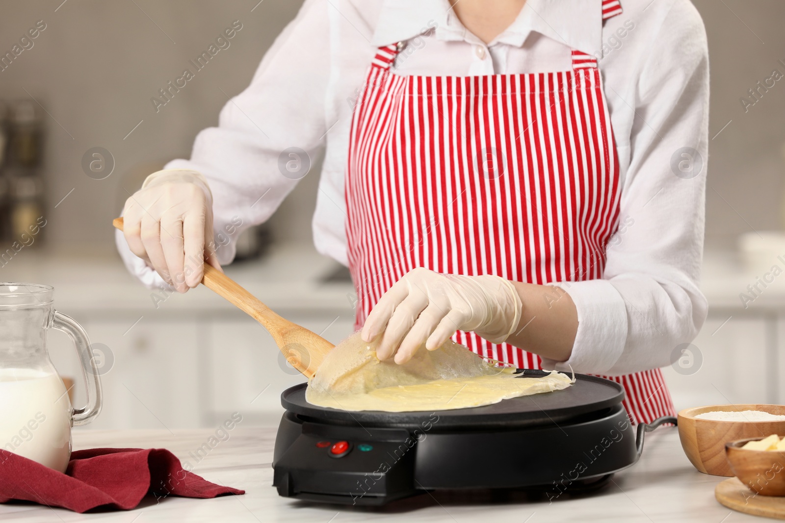 Photo of Woman cooking delicious crepe on electric pancake maker at white marble table in kitchen, closeup