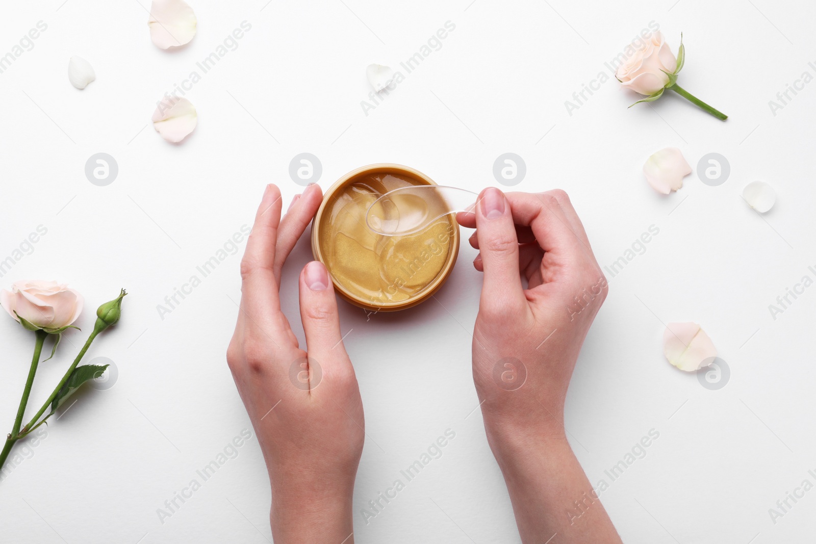 Photo of Woman taking under eye patch with spatula out of jar near rose flowers on white background, top view. Cosmetic product