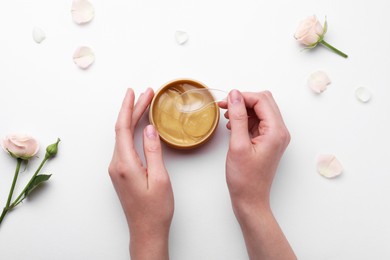 Woman taking under eye patch with spatula out of jar near rose flowers on white background, top view. Cosmetic product