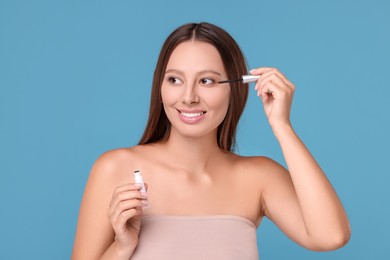 Beautiful woman applying serum onto eyelashes on light blue background