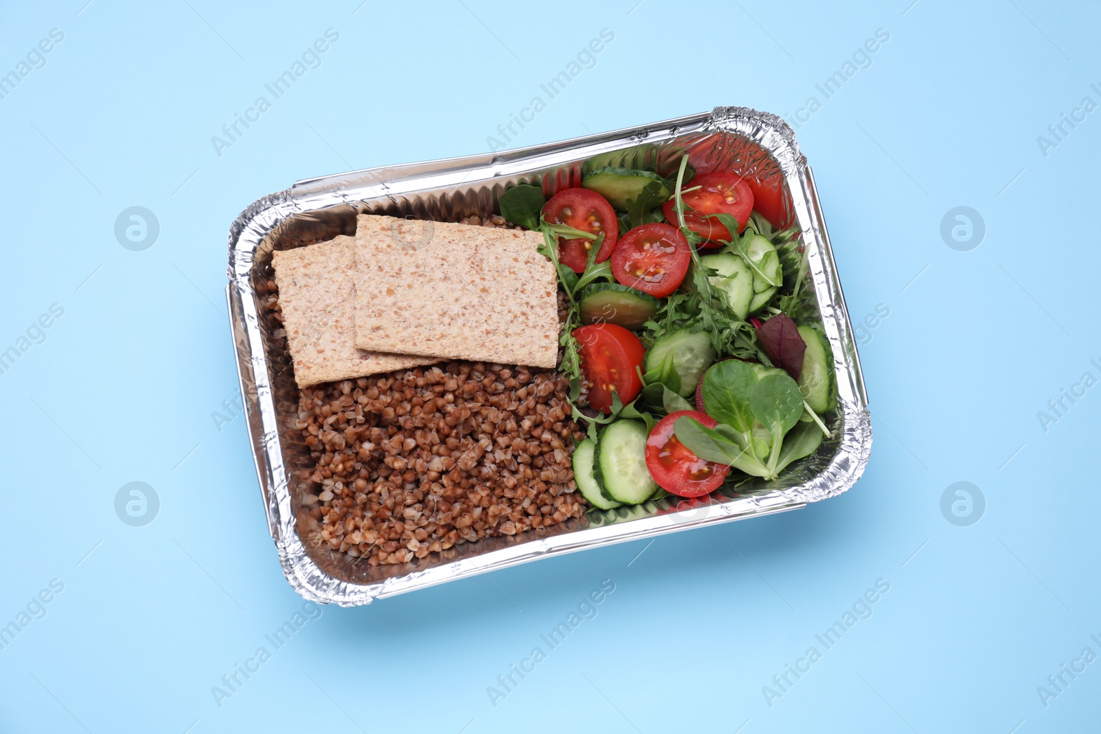 Photo of Container with buckwheat, fresh salad and crispbreads on light blue background, top view