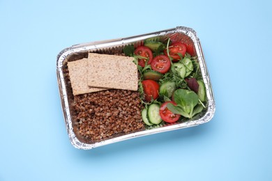 Photo of Container with buckwheat, fresh salad and crispbreads on light blue background, top view