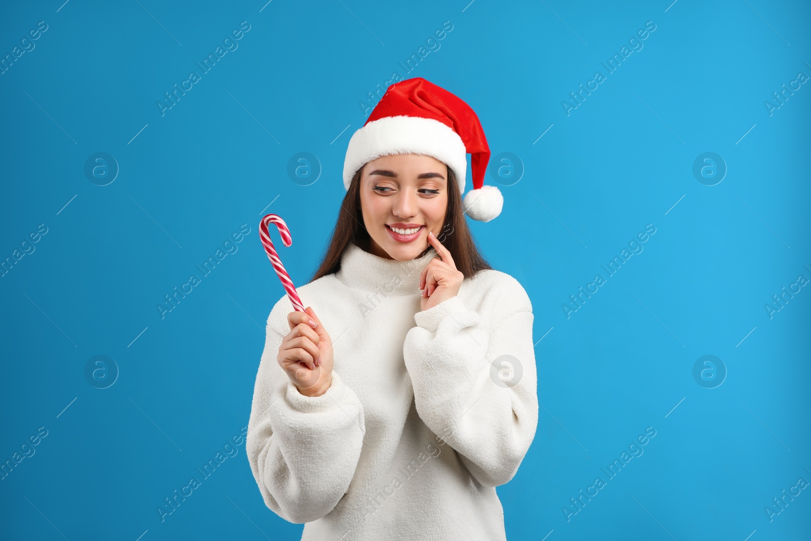 Photo of Young woman in beige sweater and Santa hat holding candy cane on blue background. Celebrating Christmas