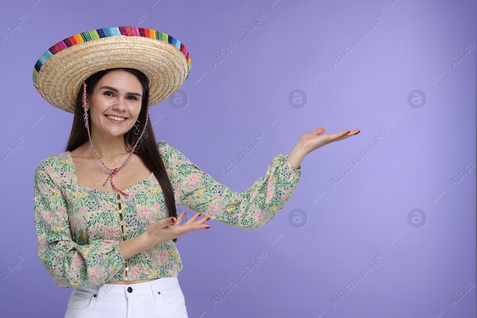 Photo of Young woman in Mexican sombrero hat showing something on violet background. Space for text