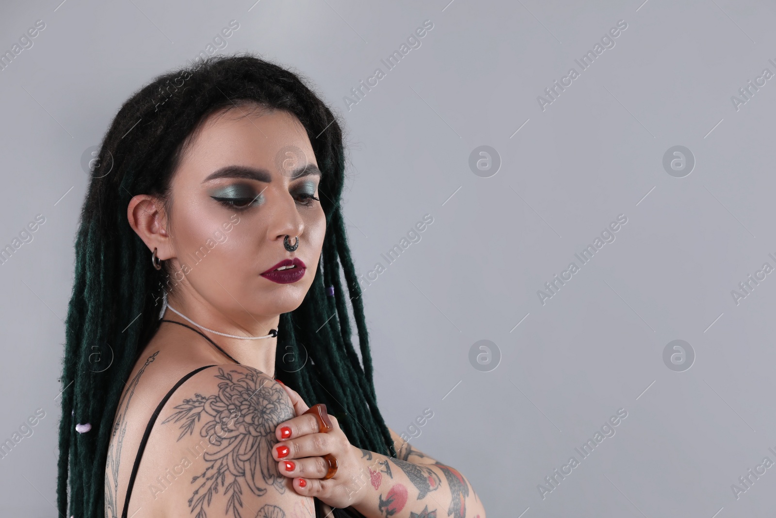 Photo of Beautiful young woman with tattoos on body, nose piercing and dreadlocks against grey background. Space for text