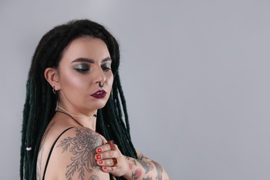 Photo of Beautiful young woman with tattoos on body, nose piercing and dreadlocks against grey background. Space for text