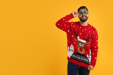 Photo of Happy young man in Christmas sweater and funny glasses on orange background. Space for text