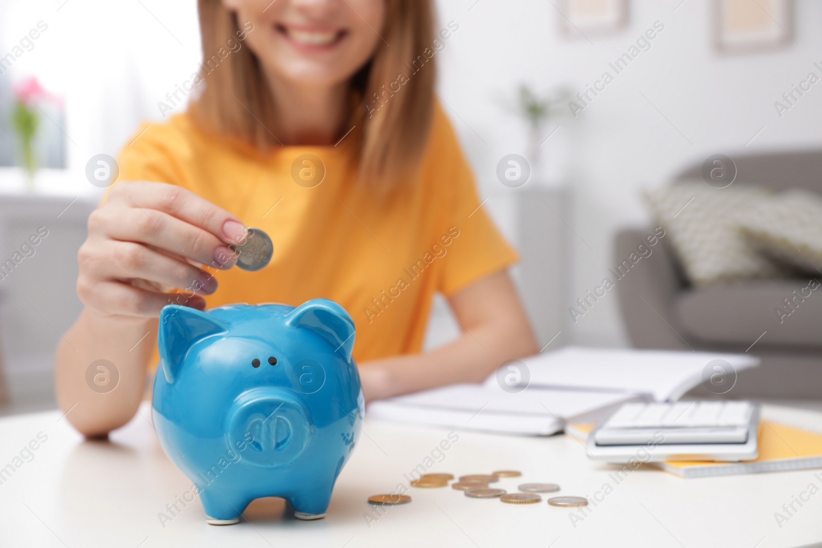 Photo of Woman putting coin into piggy bank at table in living room, closeup. Saving money