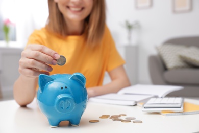 Woman putting coin into piggy bank at table in living room, closeup. Saving money