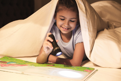 Photo of Little girl with flashlight reading book under blanket at home