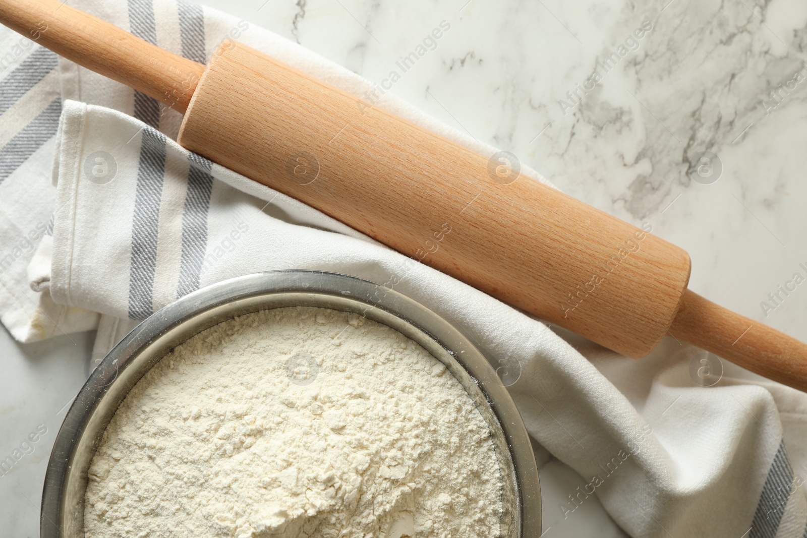 Photo of Flour in bowl, rolling pin and napkin on white marble table, top view