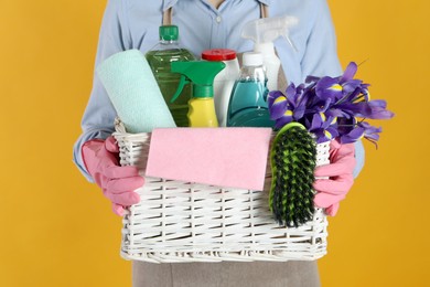 Photo of Spring cleaning. Woman holding basket with detergents, flowers and tools on orange background, closeup