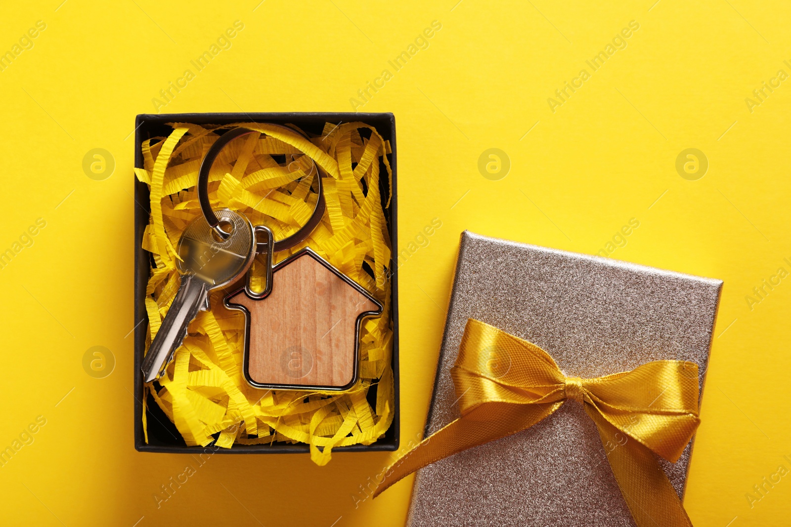 Photo of Key with trinket in shape of house and gift box on yellow background, flat lay. Housewarming party