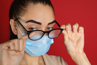 Photo of Woman wiping foggy glasses caused by wearing medical mask on red background, closeup