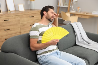 Photo of Bearded man waving yellow hand fan to cool himself on sofa at home