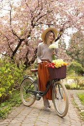 Beautiful young woman with bicycle and flowers in park on pleasant spring day