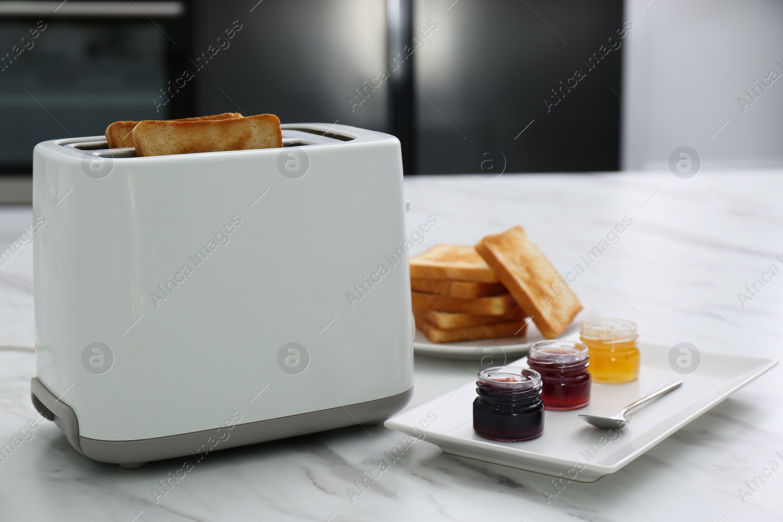 Photo of Modern toaster with slices of bread and different jams  on white marble table in kitchen