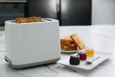 Photo of Modern toaster with slices of bread and different jams  on white marble table in kitchen
