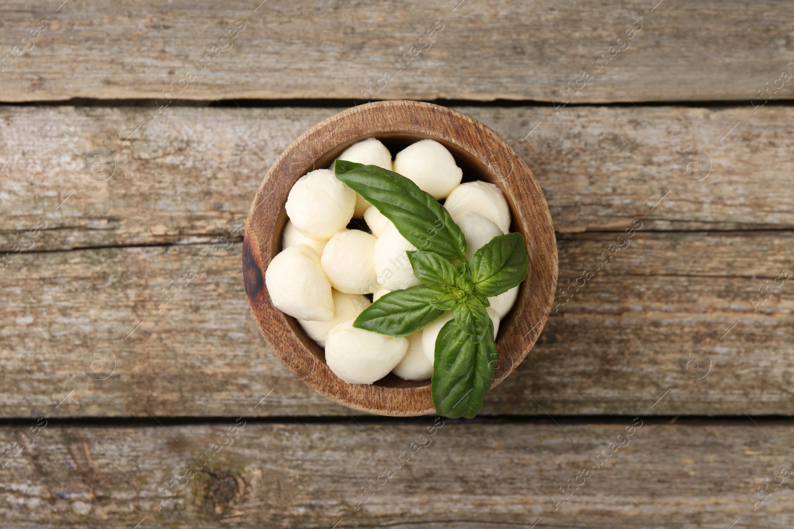 Photo of Tasty mozzarella balls and basil leaves in bowl on wooden table, top view