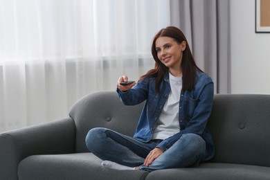 Photo of Happy woman changing TV channels with remote control on sofa at home