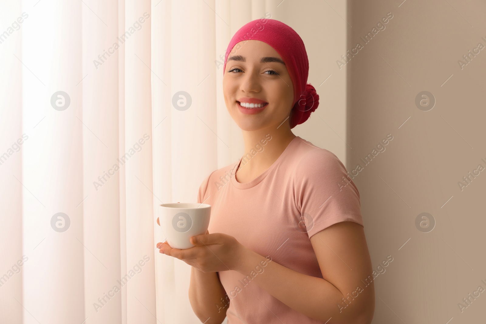 Photo of Cancer patient. Young woman with headscarf and hot drink near window indoors
