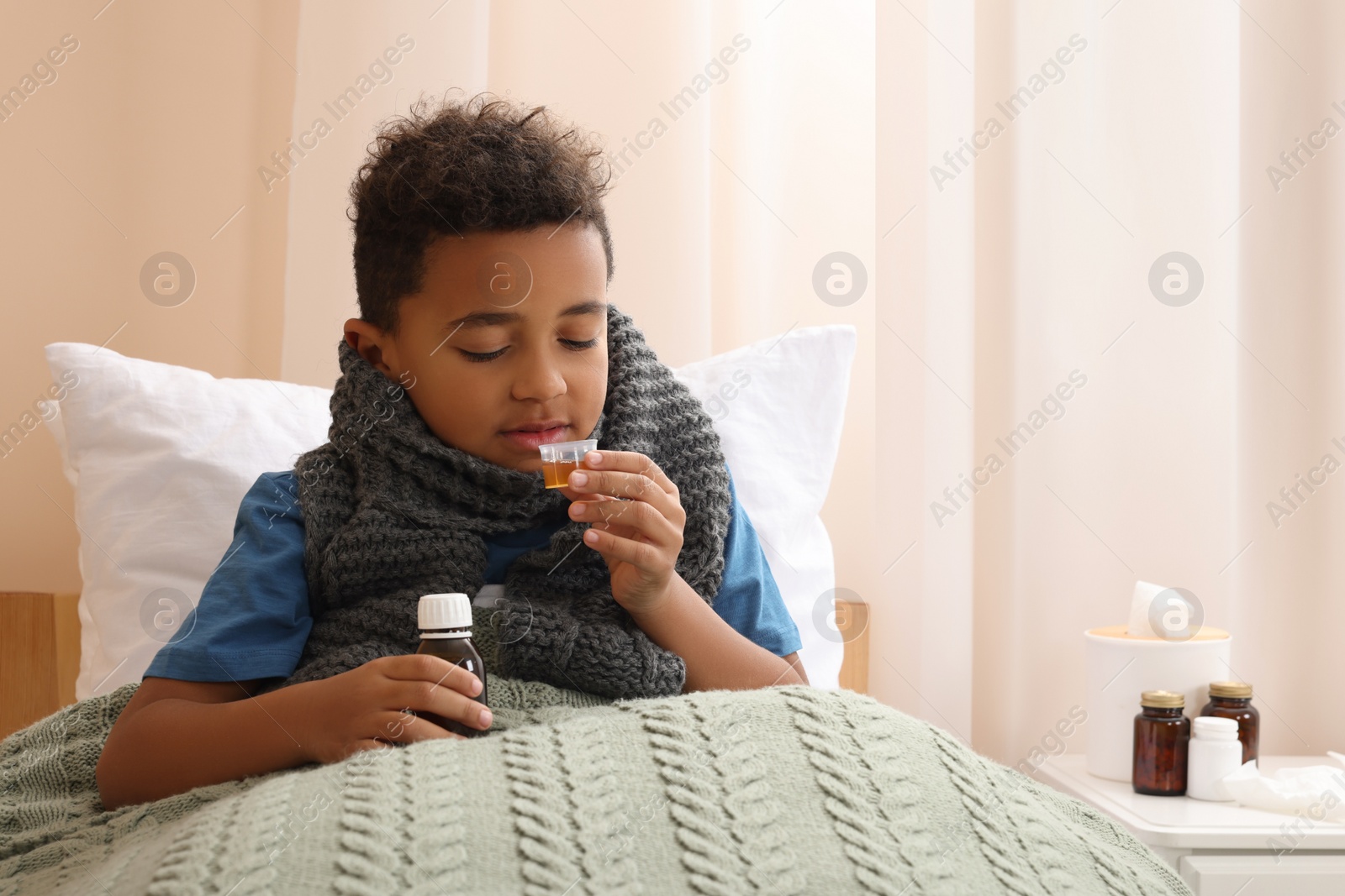 Photo of African-American boy taking cough syrup on bed at home, space for text. Cold medicine