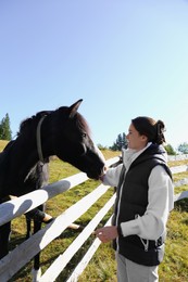 Photo of Woman stroking black horse near wooden fence outdoors. Beautiful pet